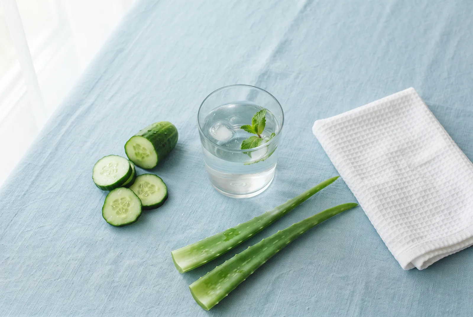 Acqua, cetriolo e foglie di aloe su tovaglia azzurra: idratazione e benessere intimo